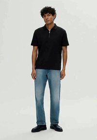 Čierna polokošeľa s krátkym rukávom a predným zipsom, skombinovaná so svetlomodrými rovnými džínami. Model má na sebe čierne topánky a stojí na neutrálnom pozadí.