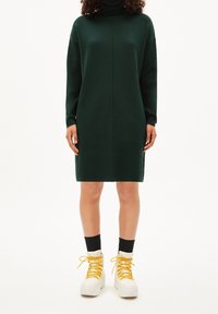 Osoba v tmavě zelených šatech s dlouhým rukávem, v černých ponožkách a bílých platformových botách se žlutými tkaničkami, stojí na jednobarevném pozadí.