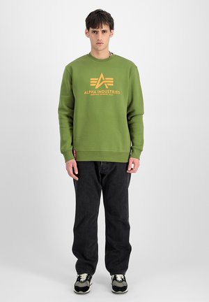 Green sweatshirt with a yellow Alpha Industries logo, crew neck, long sleeves, paired with black jeans and black sneakers with white accents.