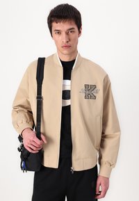 Beige bomber jacket with a zip front, ribbed collar, embroidered 'K' logo, and side pockets. Worn over a black shirt, with a black bag.