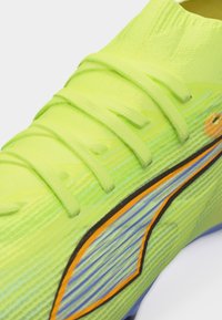 Φωτεινό κίτρινο-πράσινο αθλητικό παπούτσι με υφασμάτινη υφή, κορδόνια σε ίδιο χρώμα και σχέδιο με πορτοκαλί και μαύρη ρίγα στο πλάι.