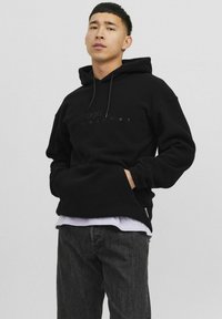 Sudadera negra de tela suave, con un bolsillo frontal y capucha con cordones. Lleva una camiseta blanca debajo y se combina con jeans oscuros.