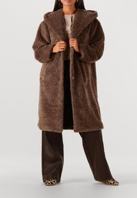 Vrouw in een lange, bruine, fluffy jas, een beige gebuttonde trui, donkere wijde broek en schoenen met luipaardprint, staand tegen een neutrale achtergrond.