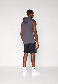 Γκρι αμάνικο φούτερ με ραφή στην πλάτη, συνδυασμένο με μαύρο αθλητικό σορτς με πλαϊνές λεπτομέρειες. Λευκά αθλητικά παπούτσια και κάλτσες ολοκληρώνουν την εμφάνιση.