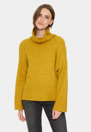 Kvinna i senapsgul polotröja och mörkgrå jeans, leende försiktigt mot en ljus vit bakgrund.