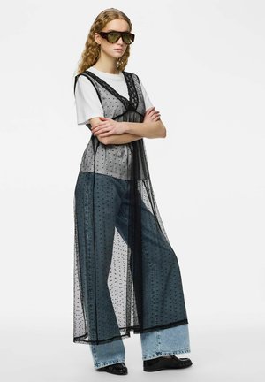 Femme portant de grandes lunettes de soleil, un t-shirt blanc, un jean large et une longue robe noire transparente à pois, debout les bras croisés.
