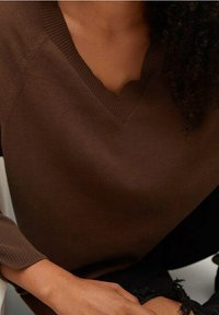 Osoba ubrana w brązowy sweter z dekoltem w serek z prążkowanymi krawędziami oraz czarne, postrzępione jeansy, siedząca z jedną ręką opartą na nodze.