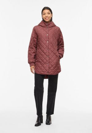 Kvinne iført en burgunder quiltet hettejakke, svarte bukser og svarte ankelstøvler, stående med hendene i jakkelommene.