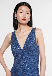 Modré flitrové šaty s hlubokým výstřihem do V, se geometrickými vzory a přiléhavým střihem. Modelka nosí náušnice ve tvaru diamantu.