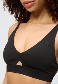 Γυναίκα που φοράει μαύρο αθλητικό σουτιέν με ένα μικρό λογότυπο κροκόδειλου στην αριστερή πλευρά και ένα V-σχήματος ανοίγματος κάτω από το στήθος.