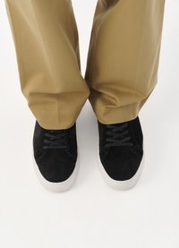 Svarte semskede sneakers med rund tå, hvit gummisåle og svarte snørehjerter, iført lysebrune bukser, som skaper en kontrast i farge og tekstur.