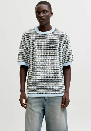 Ung mand iført en lyseblå og sort stribet strikket kortærmet sweater og falmede, løse blå jeans mod en ensartet baggrund.