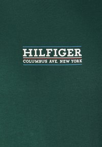 T-shirt in cotone verde con logo "HILFIGER" in bianco e blu, e "COLUMBUS AVE. NEW YORK" in accenti bianchi e rossi. Texture liscia.