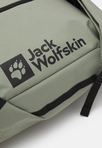 Primo piano di una borsa Jack Wolfskin verde chiaro con logo nero, impronta di zampa e una cinghia orizzontale nera su sfondo bianco.