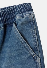 Modré denimové kalhoty s elastickým pasem. Viditelné prošívané švy a detail přední kapsy. Měkká textura, klasický rovný střih.