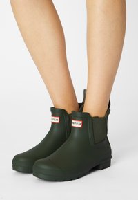 Bottes de cheville en caoutchouc vert avec des panneaux latéraux élastiques, semelles texturées et une étiquette logo rouge et blanche à l'avant. Fini lisse.