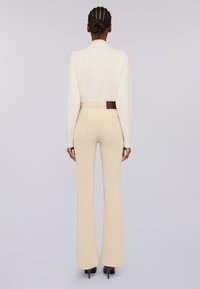 Persona con el cabello trenzado que lleva un suéter de cuello alto color crema y pantalones acampanados de tiro alto beige, de pie de espaldas frente a un fondo liso.