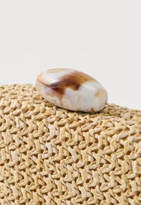 Λείο οβάλ πέτρωμα με καφέ και λευκά μαρμαροειδή σχέδια που ακουμπά σε υφασμένο μπεζ υπόστρωμα με υφή, μπροστά σε ένα απλό φόντο.