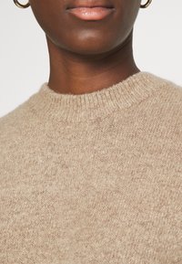 Maglione lavorato a maglia beige con scollatura rotonda, tessuto testurizzato e polsini a coste. Il materiale appare morbido e leggermente peloso.