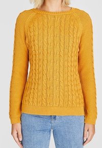 Pull en maille jaune moutarde avec motifs torsadés et texturés, col rond, et ourlets côtelés aux poignets et à l'ourlet. Porté avec un jean bleu.