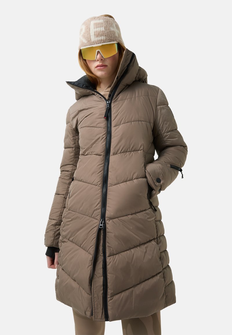 Braune Steppjacke mit Kapuze, mit vertikalem Steppmuster, Reißverschluss vorne und schwarzen Akzenten. Das Modell trägt eine Sonnenbrille und einen hellen Hut.