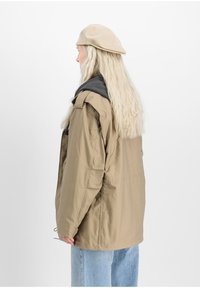 Tan outerwear jacket with a black hood, paired with light blue denim jeans. The model wears a beige beret and has long wavy hair.