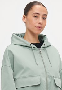 Veste à capuche vert clair avec une fermeture éclair à l'avant, deux grandes poches et des cordons de serrage ton sur ton. Texture lisse, coupe décontractée et design minimal.