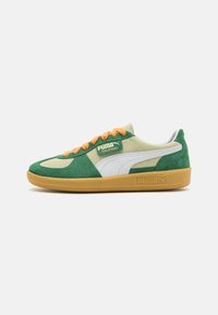 Sneaker verde e beige realizzata con materiali in suede e pelle, dotata di lacci arancioni, una striscia laterale bianca e una suola in gomma. Etichetta "PUMA PALERMO" presente.