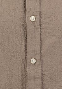 Bruine gestructureerde shirt met een knopenlijst met twee witte ronde knopen, licht gekreukt stof en een zichtbare stikseldetail langs de rand.