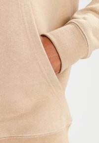 Felpa beige con tasca a marsupio, polsini a costine e una texture morbida. Cuciture visibili e dettagli di vestibilità rilassata.