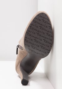 Botte à talon en daim beige avec fermeture éclair latérale, bout pointu et semelle en caoutchouc noire texturée portant l'inscription du nom de la marque. Taille indiquée : 37.