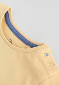 Cuello de prenda infantil amarilla con ribete interior azul y un botón a presión amarillo decorado con una flor azul en el hombro.