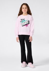 Chica con cabello largo y rizado, vistiendo un sudadera rosa con el texto "NYC Vibes? YASS!", pantalones negros y zapatillas rosas, de pie frente a un fondo blanco.