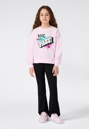 Chica con cabello largo y rizado, vistiendo un sudadera rosa con el texto "NYC Vibes? YASS!", pantalones negros y zapatillas rosas, de pie frente a un fondo blanco.