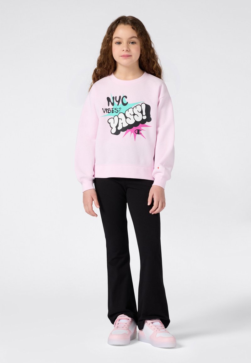 Chica con cabello largo y rizado, vistiendo un sudadera rosa con el texto "NYC Vibes? YASS!", pantalones negros y zapatillas rosas, de pie frente a un fondo blanco.