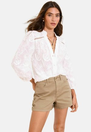 Kvinde iført hvid langærmet broderet bluse med pufærmer og beige højtaljede shorts med snøre-detaljer i siden.