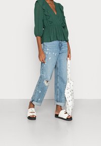 Top verde testurizzato con dettagli arricciati, jeans blu strappati con macchie di vernice e sandali bianchi con cinturino. Borsa a tracolla con motivi neri.