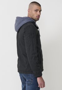 Melnā jaka ar pelēku kapuci, ar plecu uzšuvi, priekšējiem kabatām un rāvējslēdzēju. Gluda auduma, brīvas piegriezuma.