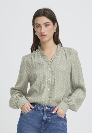 Donna che indossa una camicetta floreale verde chiaro con colletto e polsini arricciati, abbinata a jeans blu, su uno sfondo semplice