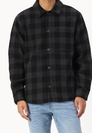 Man draagt een zwart-grijs ruitjes overhemd met borstzak en lichtblauwe spijkerbroek, staand tegen een witte achtergrond.