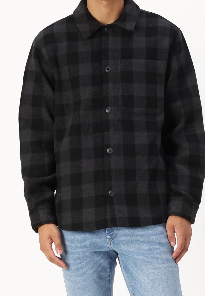 Man wearing black and dark gray checkered button-up shirt with chest pocket and light blue jeans, standing against white background.