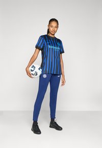 Melna un zila svītraina futbola krekls ar logo, apvienots ar tumši zilām sporta legingām un melniem futbola apaviem. Tur rokas futbola bumbu.