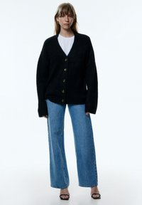 Schwarze gestrickte Strickjacke mit tiefem V-Ausschnitt, fünf goldenen Knöpfen auf der Vorderseite und übergroßen Ärmeln, getragen über einem weißen Oberteil und blauen Jeans.