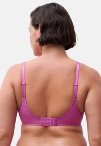 Frau mit kurzem dunklem Haar, die einen pinken BH mit verstellbaren Trägern und Haken-Ösen-Verschluss am Rücken trägt, von hinten vor weißem Hintergrund gezeigt.