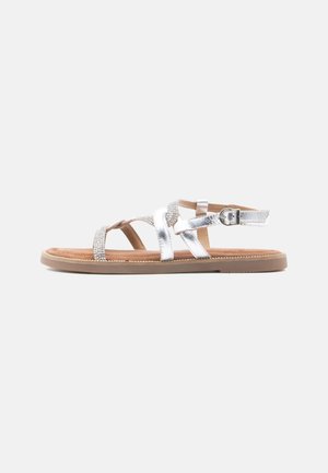 Silberne metallische Sandalen mit Kreuzriemen und Strass-Akzenten, flacher Sohle und verstellbarem Knöchelverschluss. Braunes strukturiertes Fußbett.