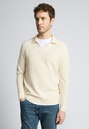 Mężczyzna o kręconych włosach, ubrany w kremowy sweter z dzianiny na białej koszuli i niebieskich dżinsach, stoi z jedną ręką w kieszeni na białym tle.