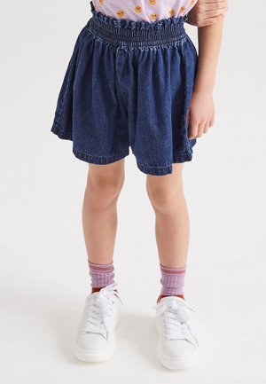 Barn iført blå denimshorts, lilla mønstrede sokker og hvide sneakers, stående på en hvid baggrund.