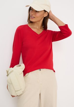 Chemise rouge à manches longues et col en V en tissu doux. Associée à un pantalon beige, la tenue comprend une casquette de couleur claire et un sac crème.