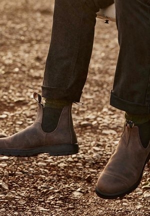 Stivali Chelsea in pelle marrone con pannelli laterali elastici, suole in gomma e linguette, abbinati a jeans scuri arrotolati e calzini verdi.
