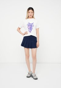 Young woman standing with hand on hip wearing white t-shirt with purple cartoon bat and navy pleated skirt, gray sandals.